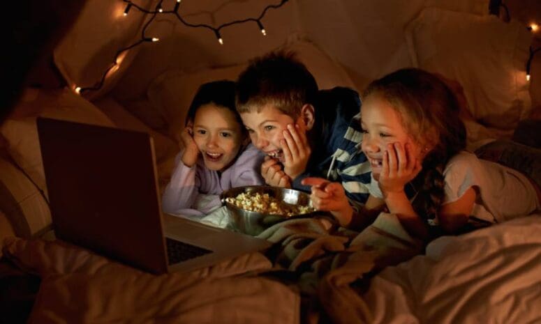 Three smiling, young kids in a blanket fort are eating popcorn and watching a movie on a laptop. String lights are hanging.