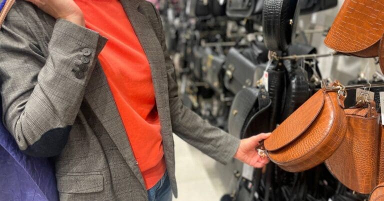 A woman in a gray business suit with shopping bags over her shoulder looks at a brown leather purse inside a store.