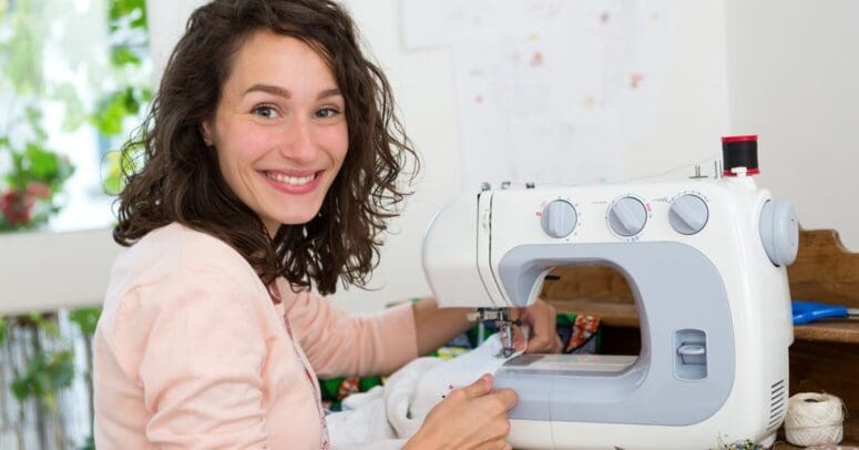 A woman smiles as she sits next to a white sewing machine. She's working a piece of white fabric through it.