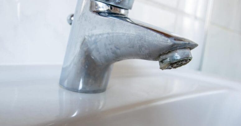 A close-up of a bathroom sink faucet, the metal dulled and blocked with years of limescale buildup.