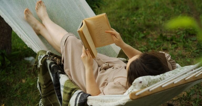 A woman lays stretched out in a hammock. A green, white, and black blanket stretches over it as she reads a book.