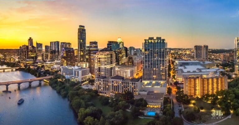 An aerial view of the skyline of Austin, Texas, at sunset. The view includes a river, bridges, and boats on the river.
