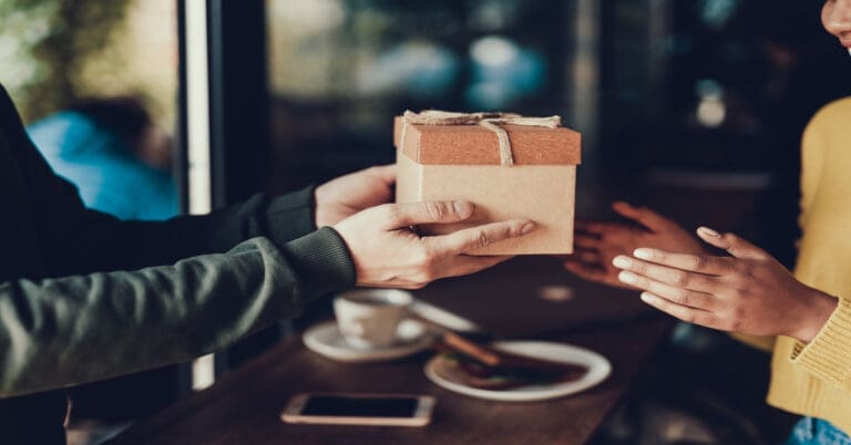 In a cafe, a woman in a green sweater hands a small gift in a brown box to a woman in a yellow sweater.