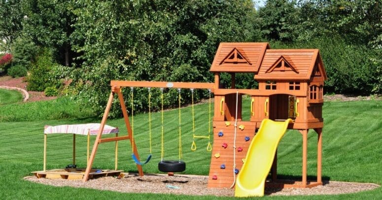 A wooden swing set is in a residential backyard. It features a blue swing, a yellow slide, a tire swing, and a covered sandbox.
