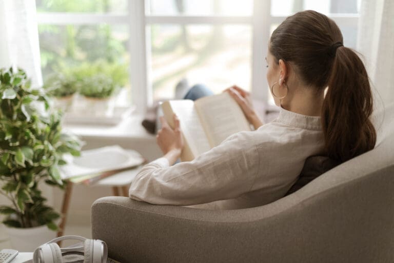 Over-the-shoulder view of a woman sitting at home and reading a book. Sunlight shines through the windows.