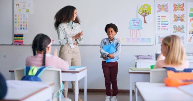 A teacher applauds a smiling boy holding a blue folder as three classmates watch from desks in a colorful classroom.