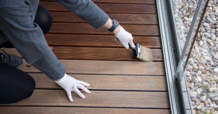 A man with white gloves kneeling to apply a layer of wood protection oil to hardwood floors next to a screen door.