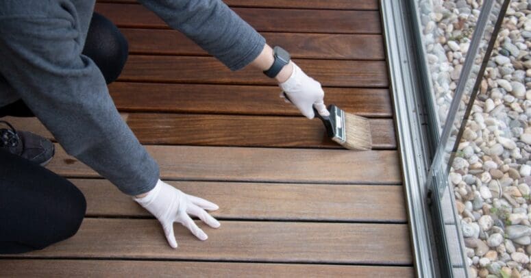 A man with white gloves kneeling to apply a layer of wood protection oil to hardwood floors next to a screen door.